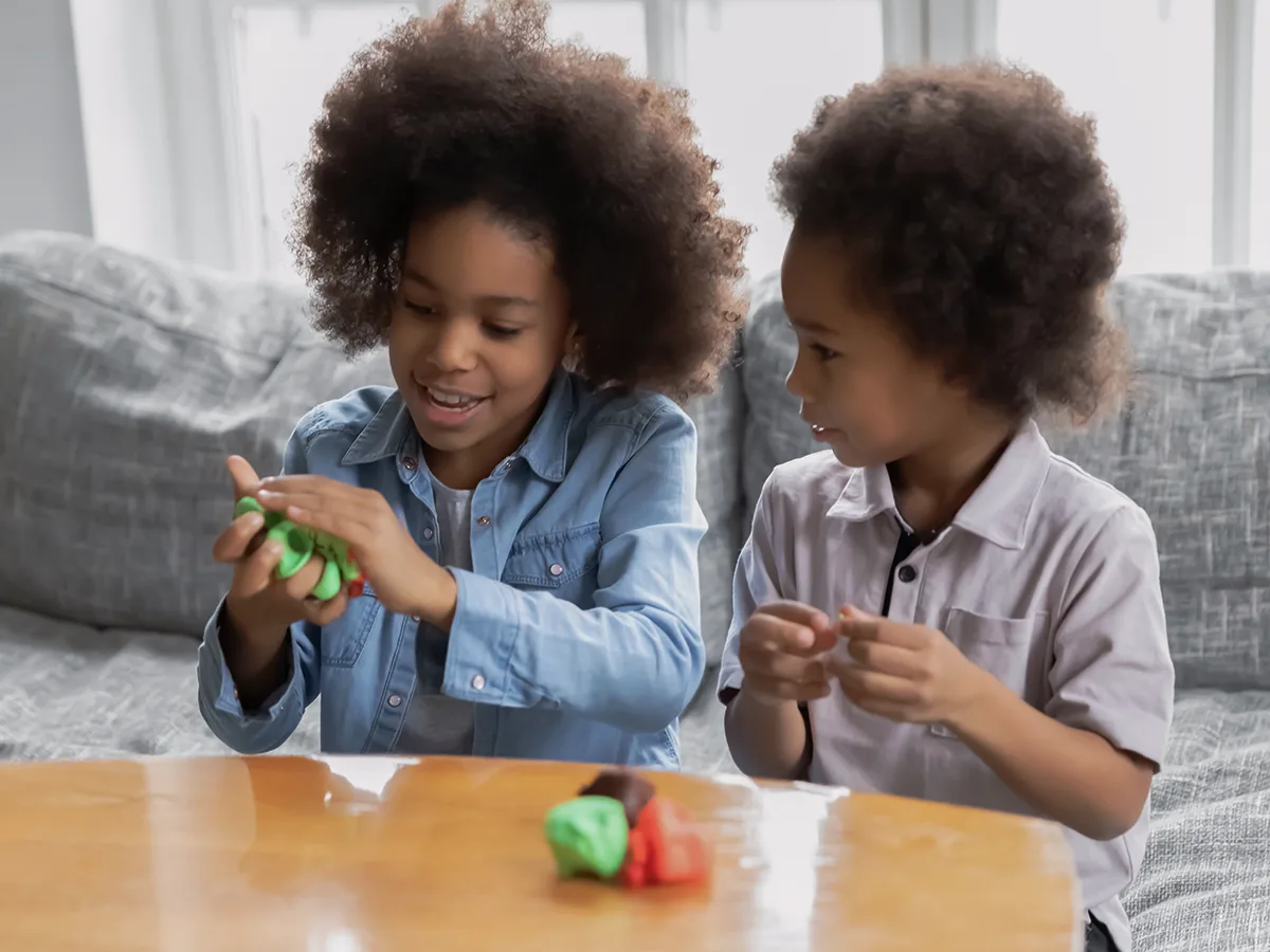 Un niño usa ambas manos para modelar plastilina. El otro lo observa, intentando imitar la posición de las manos. Su plastilina está sobre la mesa.