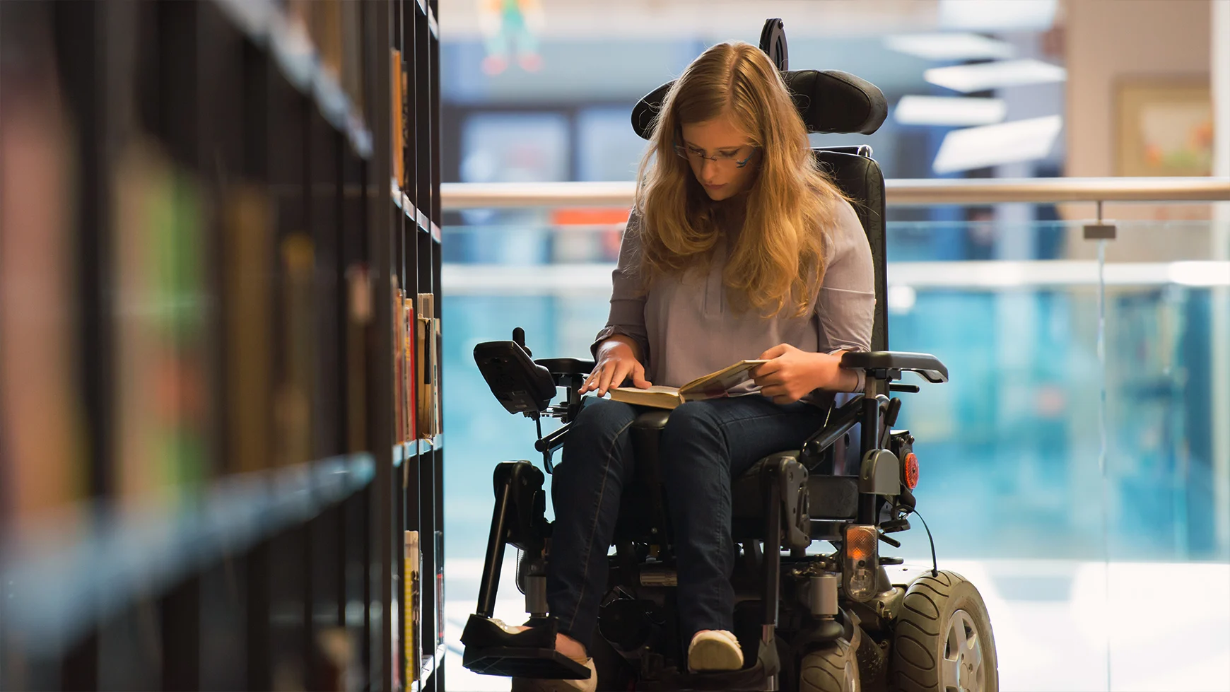 A child with long hair and glasses is holding and reading a book at the library while sitting in their wheelchair.