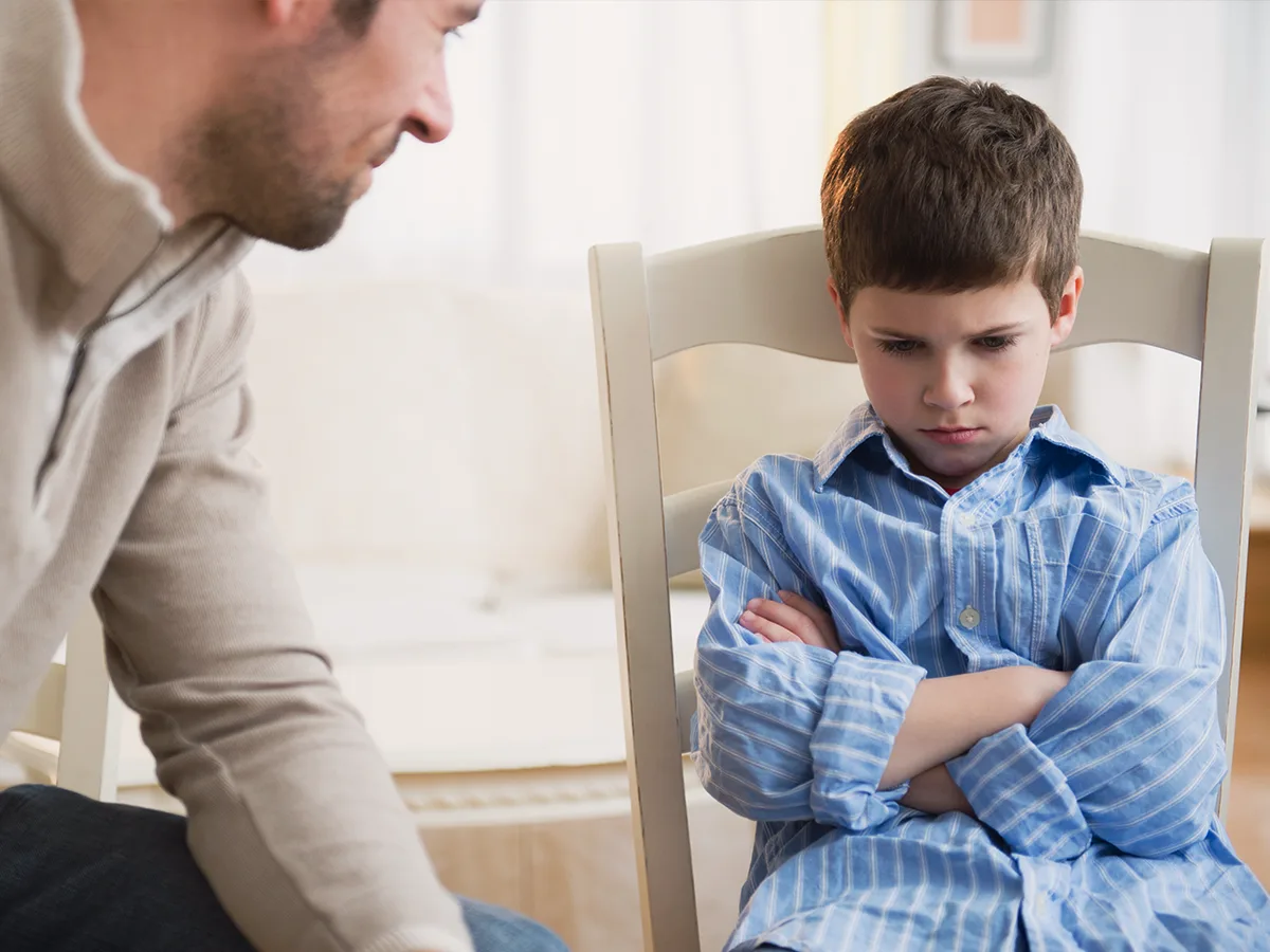 ADHD, anxiety, and anger: how they're linked, angry little boy sitting slouched in a chair with arms crossed, father crouched down next to him, looking at him 
