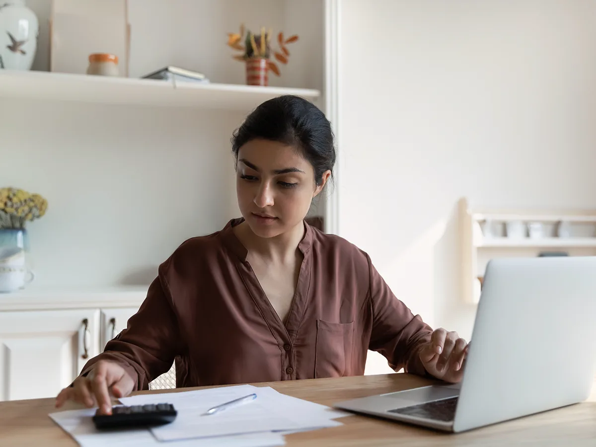 An adult types into a calculator at a desk with a laptop, paper, and pen on it.