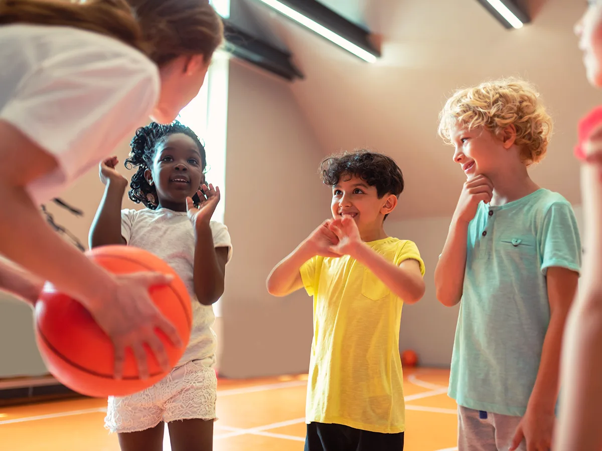 How to get your child involved in sports, Woman leaning over holding a basketball on a basketball court next to a young girl standing holder her arms in the air, a boy standing and holding his arms up in front of his face, and a boy standing holding one arm under his chin. 