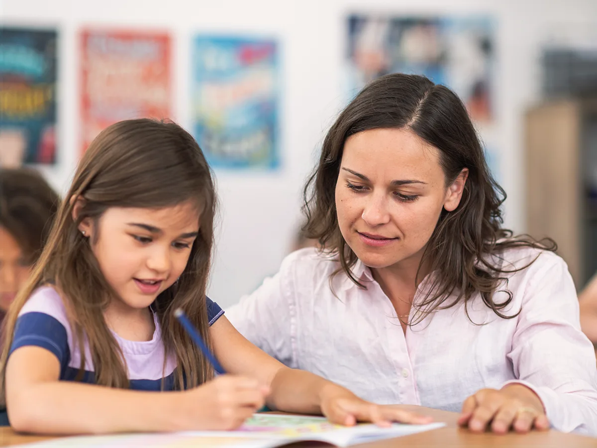 A child writes in a workbook while sitting in a classroom. An adult is sits nearby.