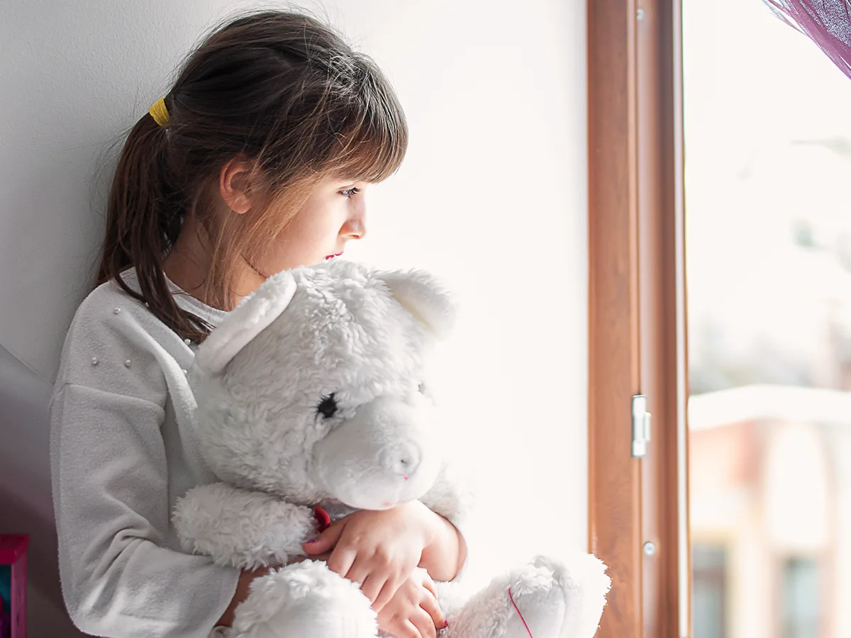 A child sitting against a wall, embracing a large white teddy bear with both arms, gazes out of a window.