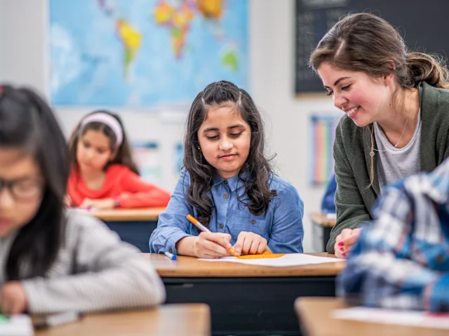 A child writes on a piece of paper at a desk in a classroom surrounded by other students. An adult stands near the child.
