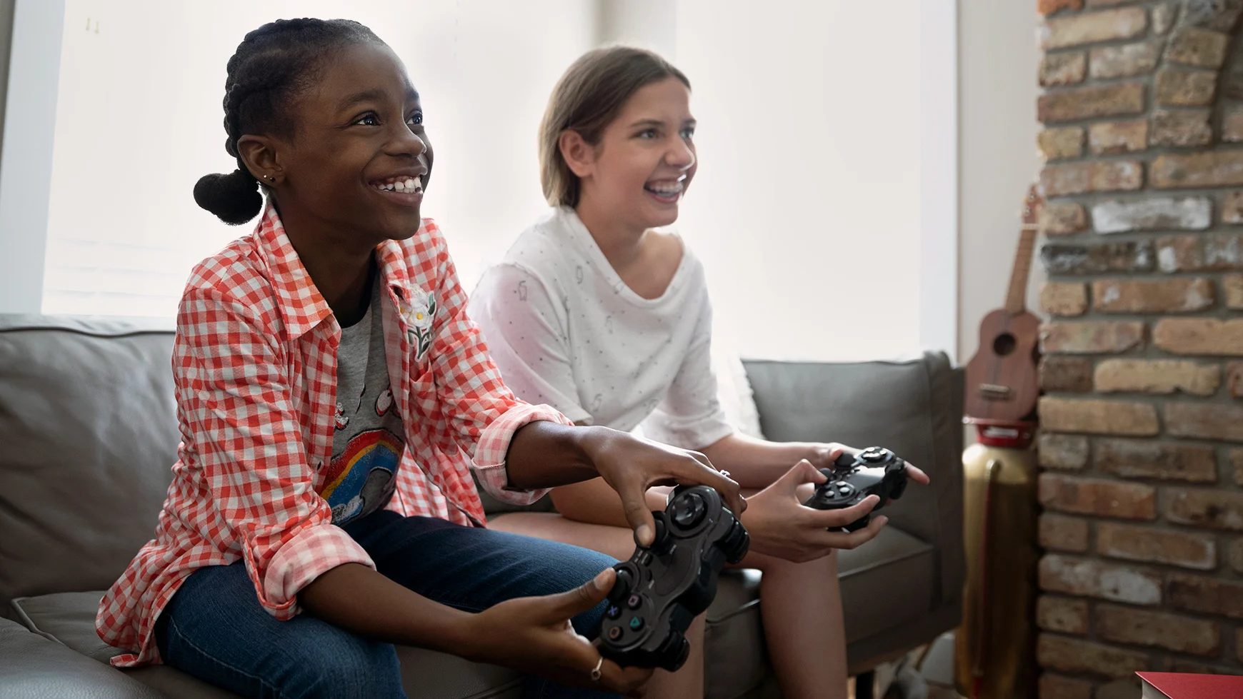 Two smiling children sit together on a sofa, playing a video game and holding controllers while watching the screen.