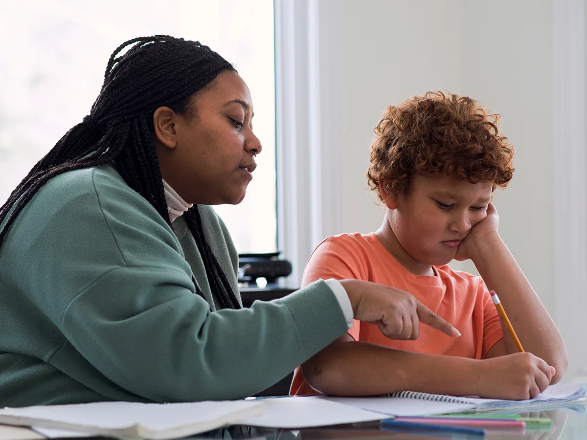 Una persona adulta ayuda a un niño con los deberes escolares. El niño sostiene su cabeza con una mano y escribe con la otra.