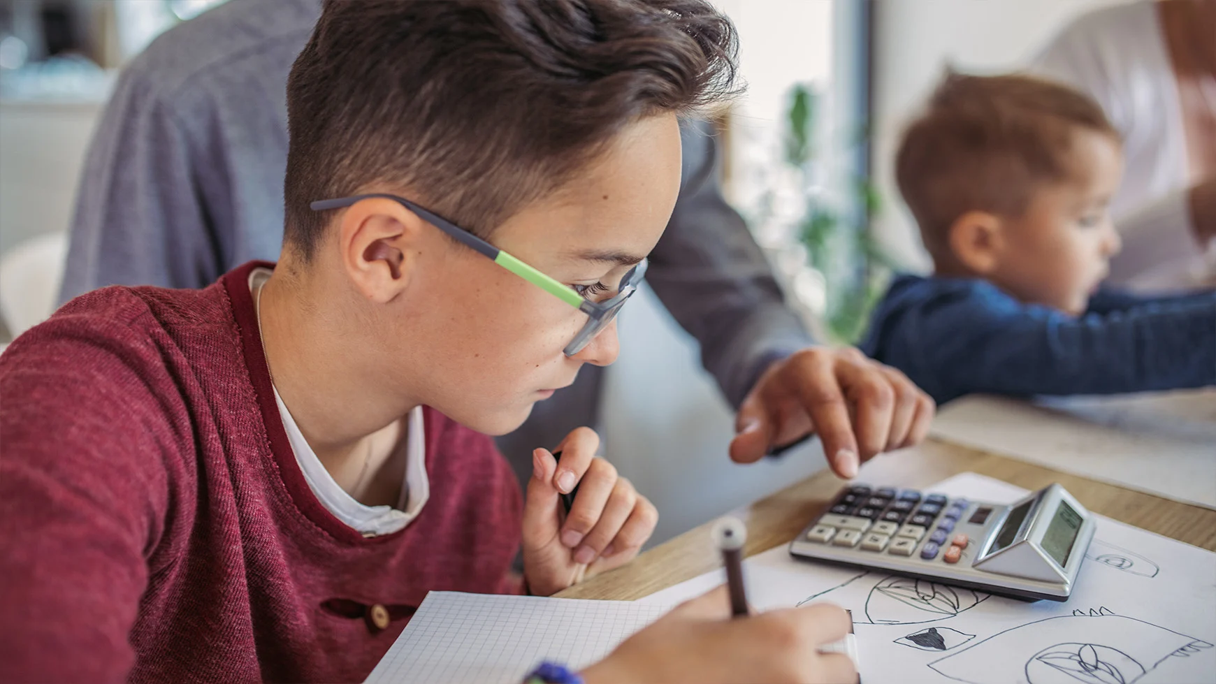 A parent uses a calculator next to their child, who wears glasses and does math homework with a pencil. 