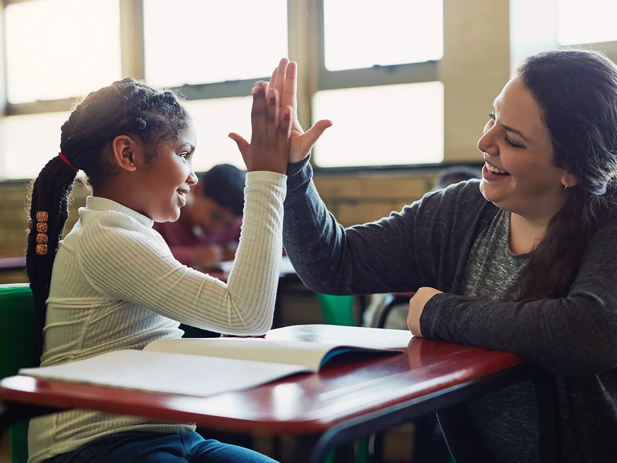 A student has a book open on their desk and is high fiving with their teacher. Both are smiling. 