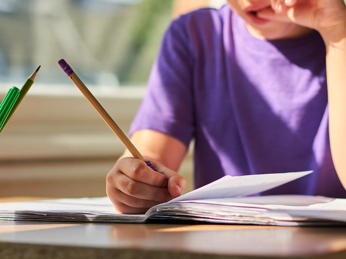 A child sits at a desk and is doing school work in a notebook with a pencil while biting the nails of their other hand. 