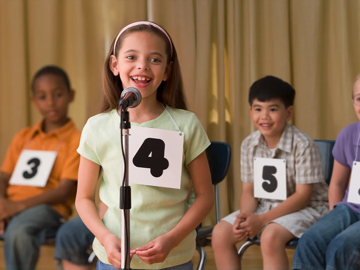 A smiling child wearing number 4 speaks into a microphone. Kids wearing numbers 3 and 5 smile from their seats behind her.

