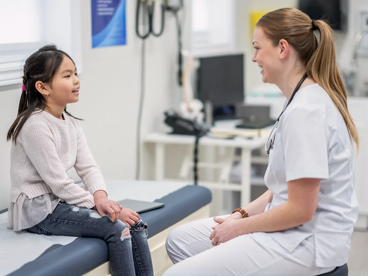 A child sits on an examination table in a doctor's office talking to a doctor. 