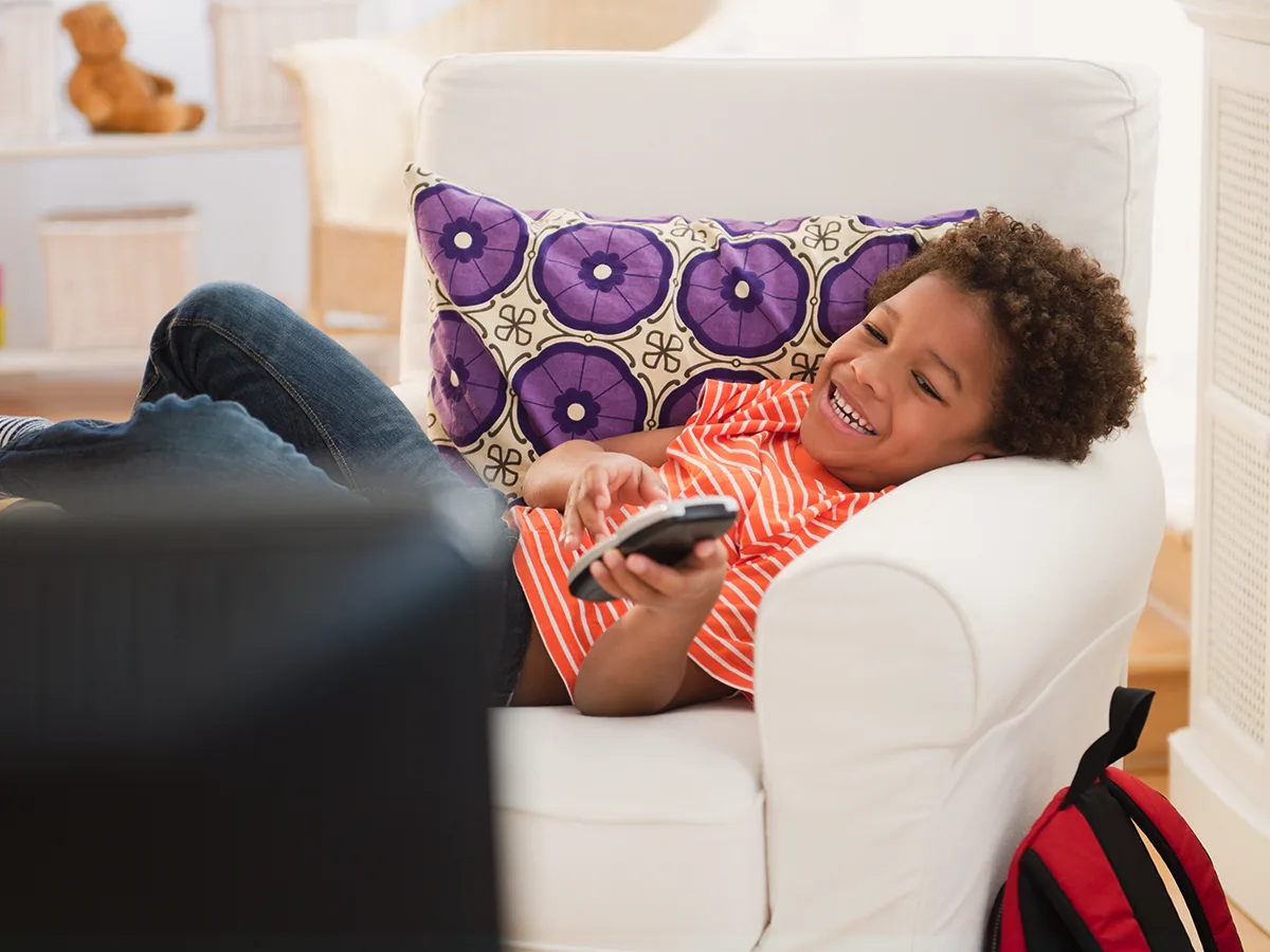 A smiling child lays on a chair with a remote watching TV. 