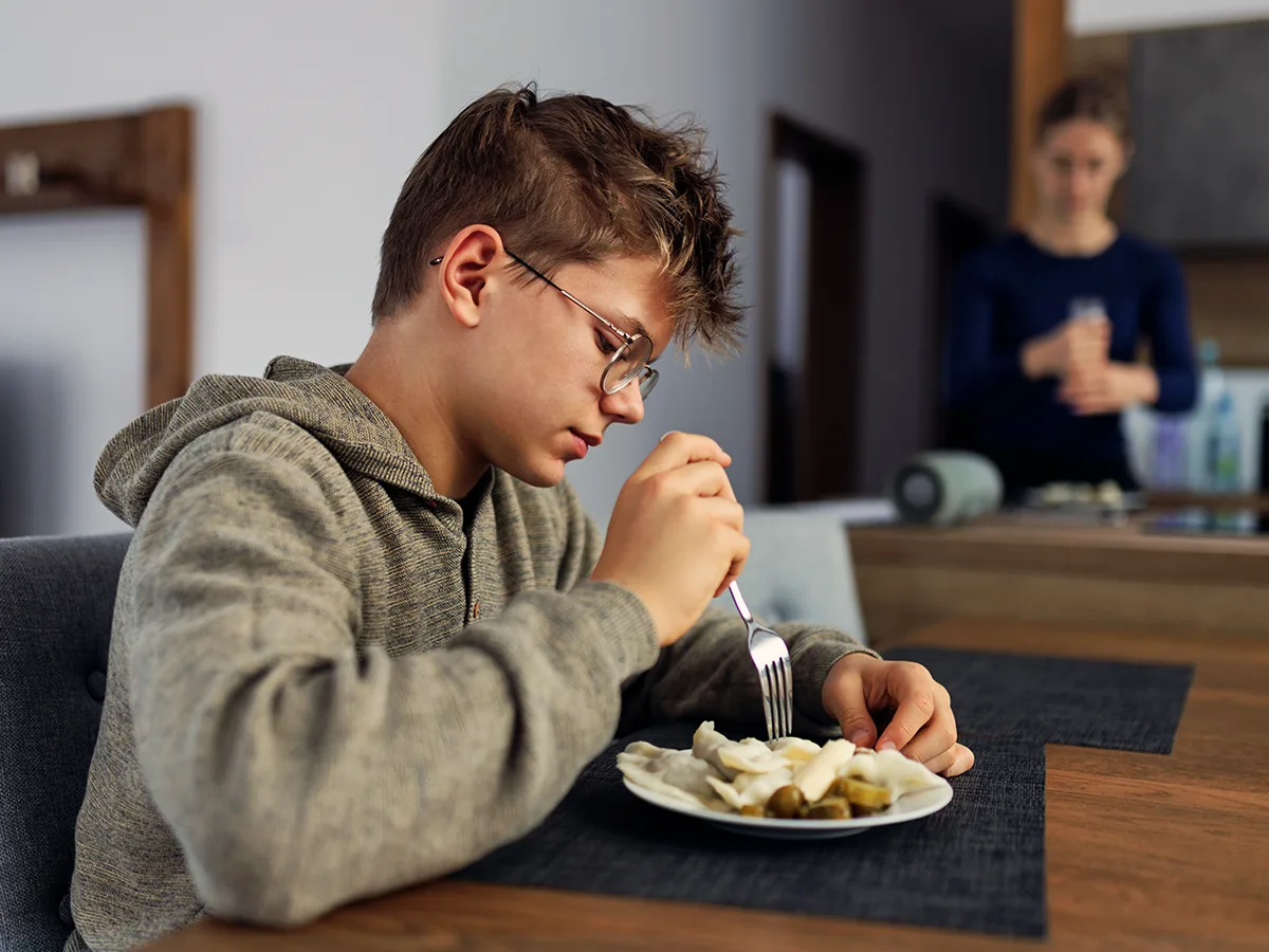 A child eats food at a kitchen table. An adult is standing in the background. 