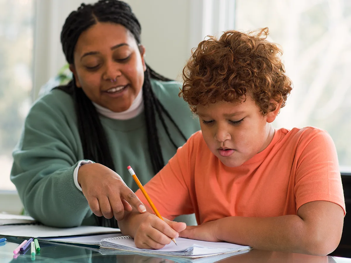 An adult sits by a child who is writing on a piece of paper. 