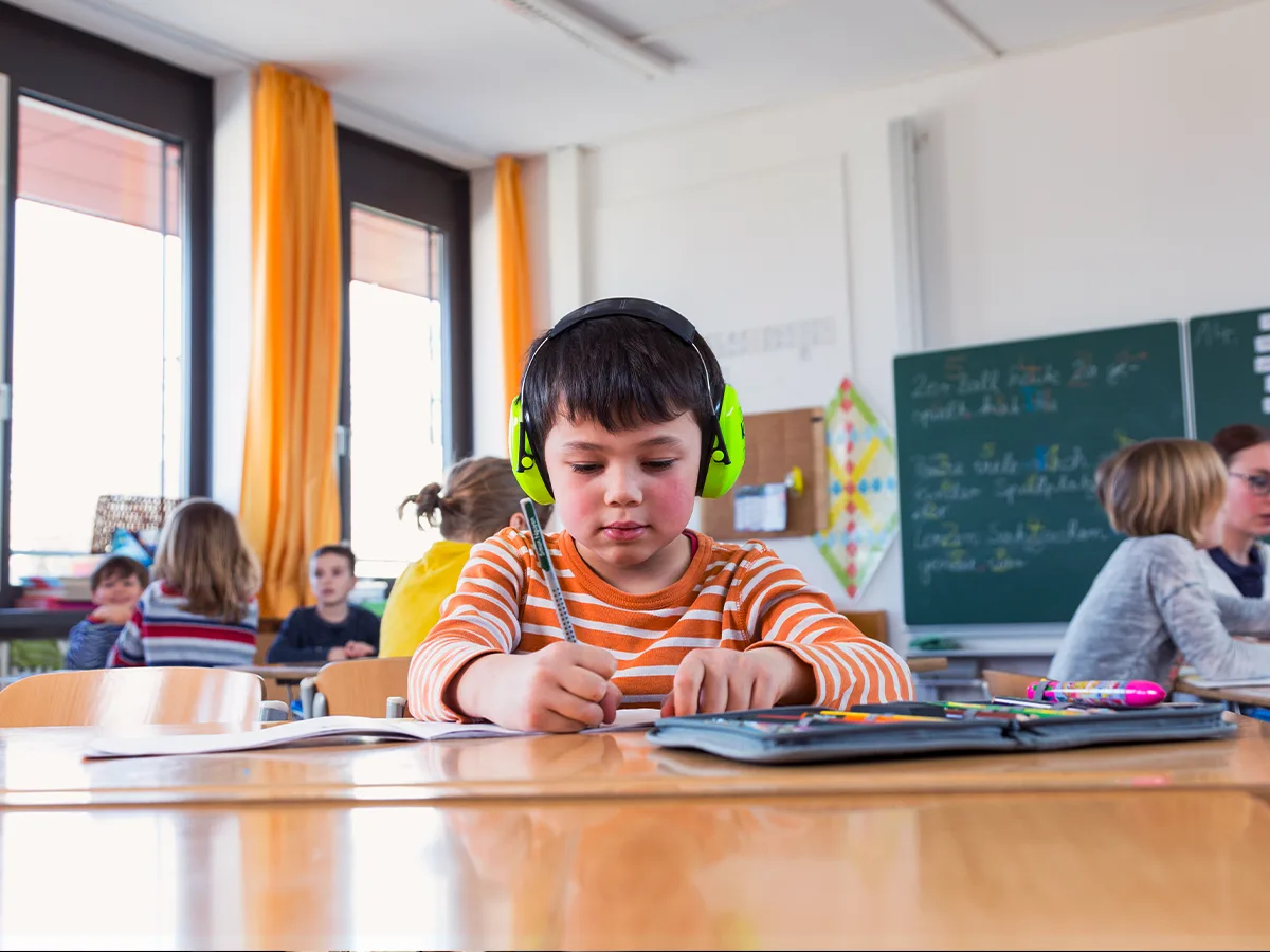 In a classroom, one of five students is wearing noise-canceling neon green headphones while writing in a notebook.