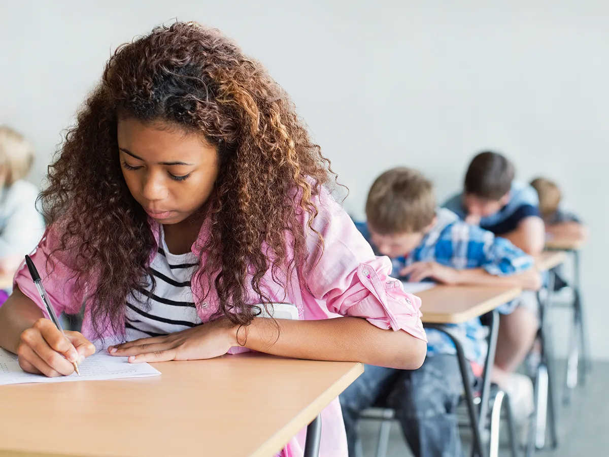 Accommodations for state standardized tests, african american girl sitting at a desk writing on a piece of paper with other kids also sitting in desks writing on paper