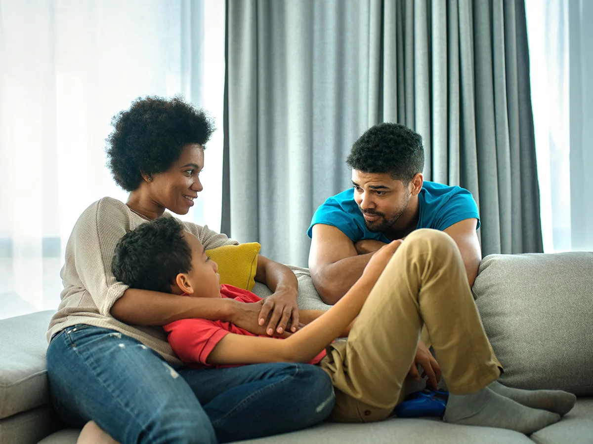 Parents and child talk in the living room. The mother hugs the child, who is relaxed, while the father leans over the sofa.