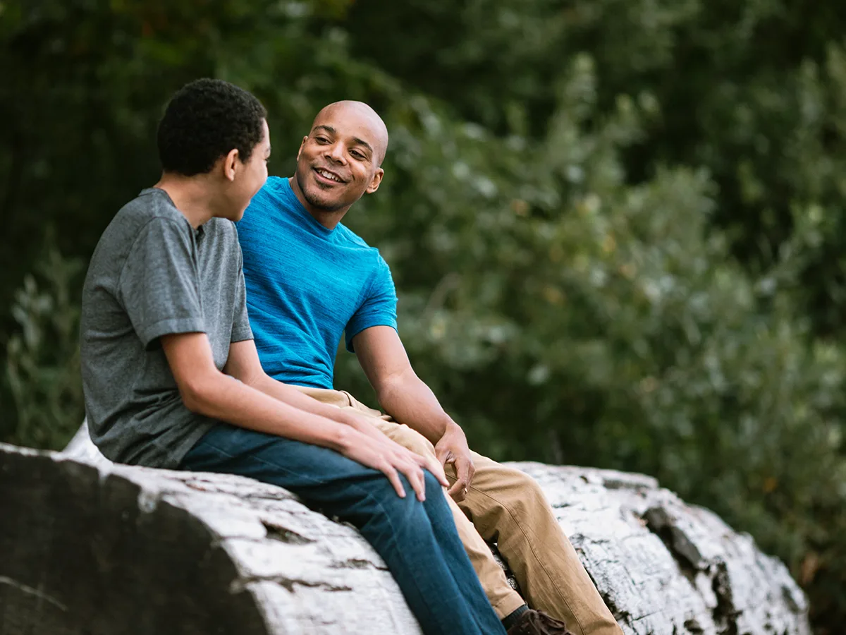 An adult and child sit outside on a large log. They are both smiling and engaged in conversation. 