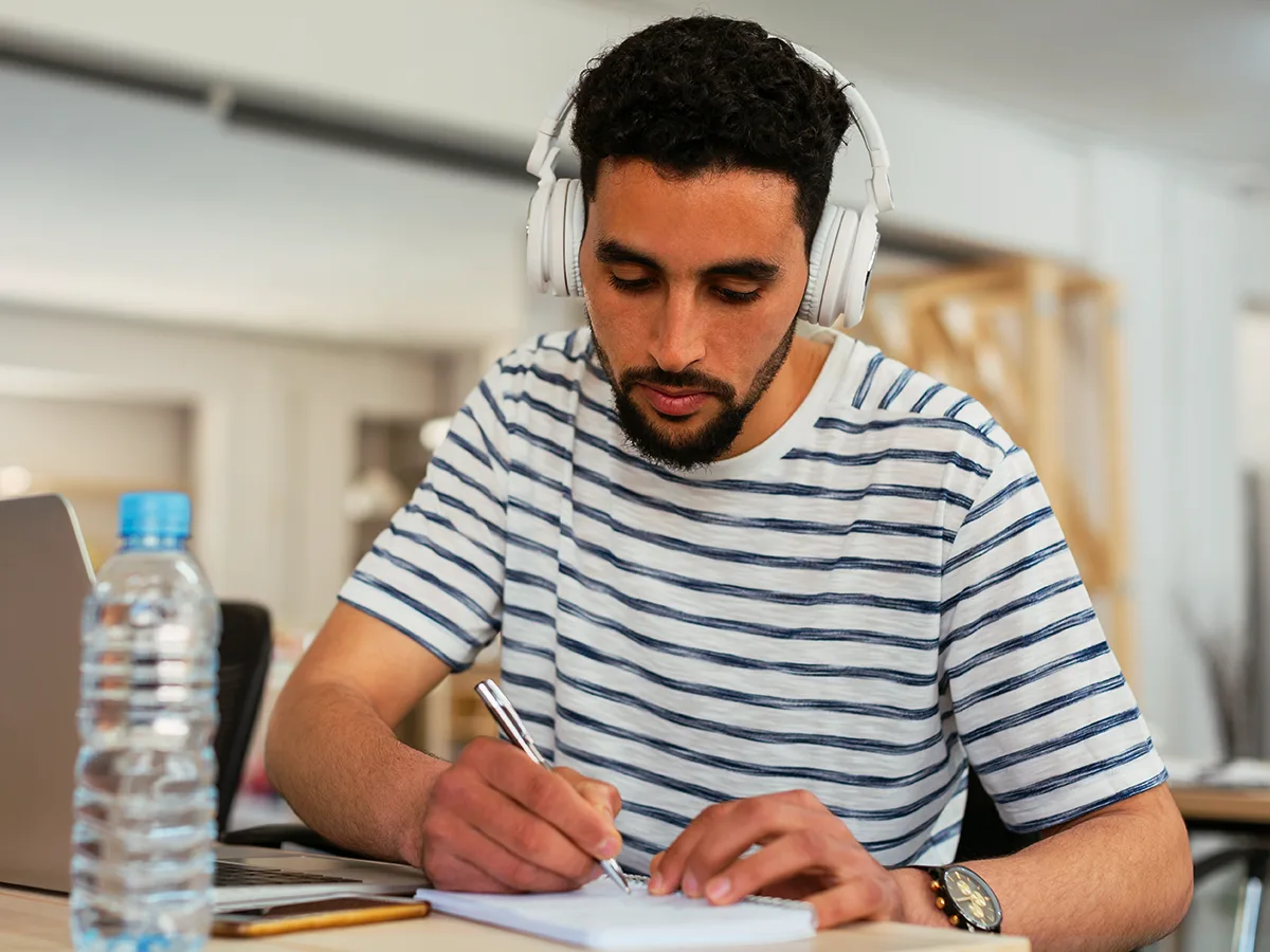 An adult wearing headphones writing on a piece of paper with a pen. 