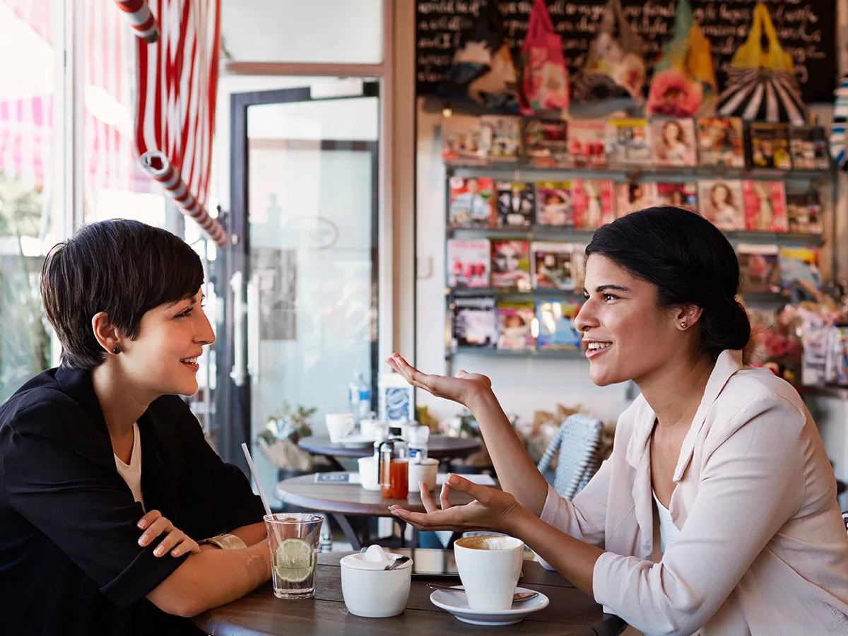Dos mujeres adultas sentadas en una mesa conversan mientras toman café. 