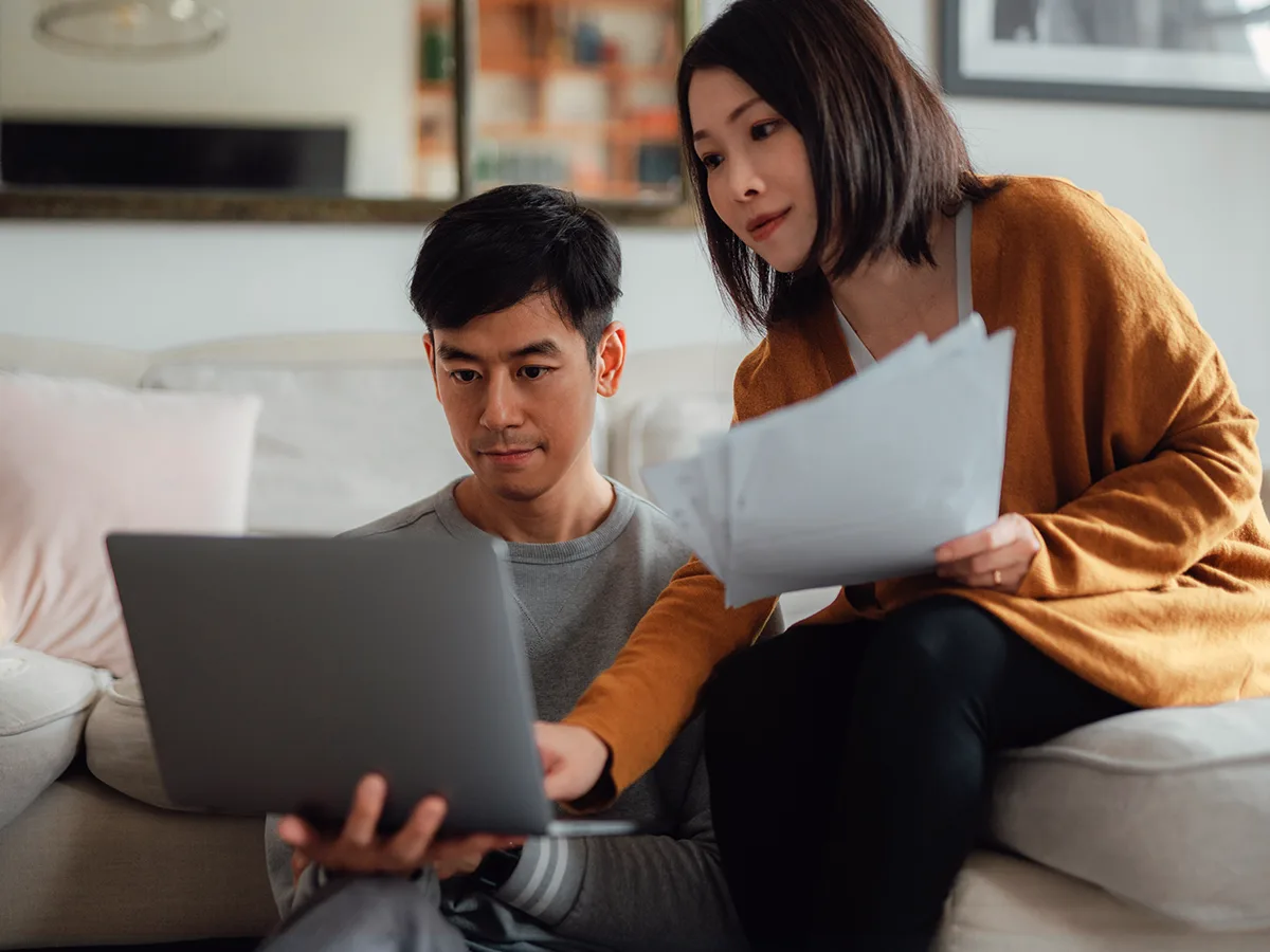 Two adults sit on a sofa. One holds a laptop while the other holds paperwork, pointing to something on the screen.