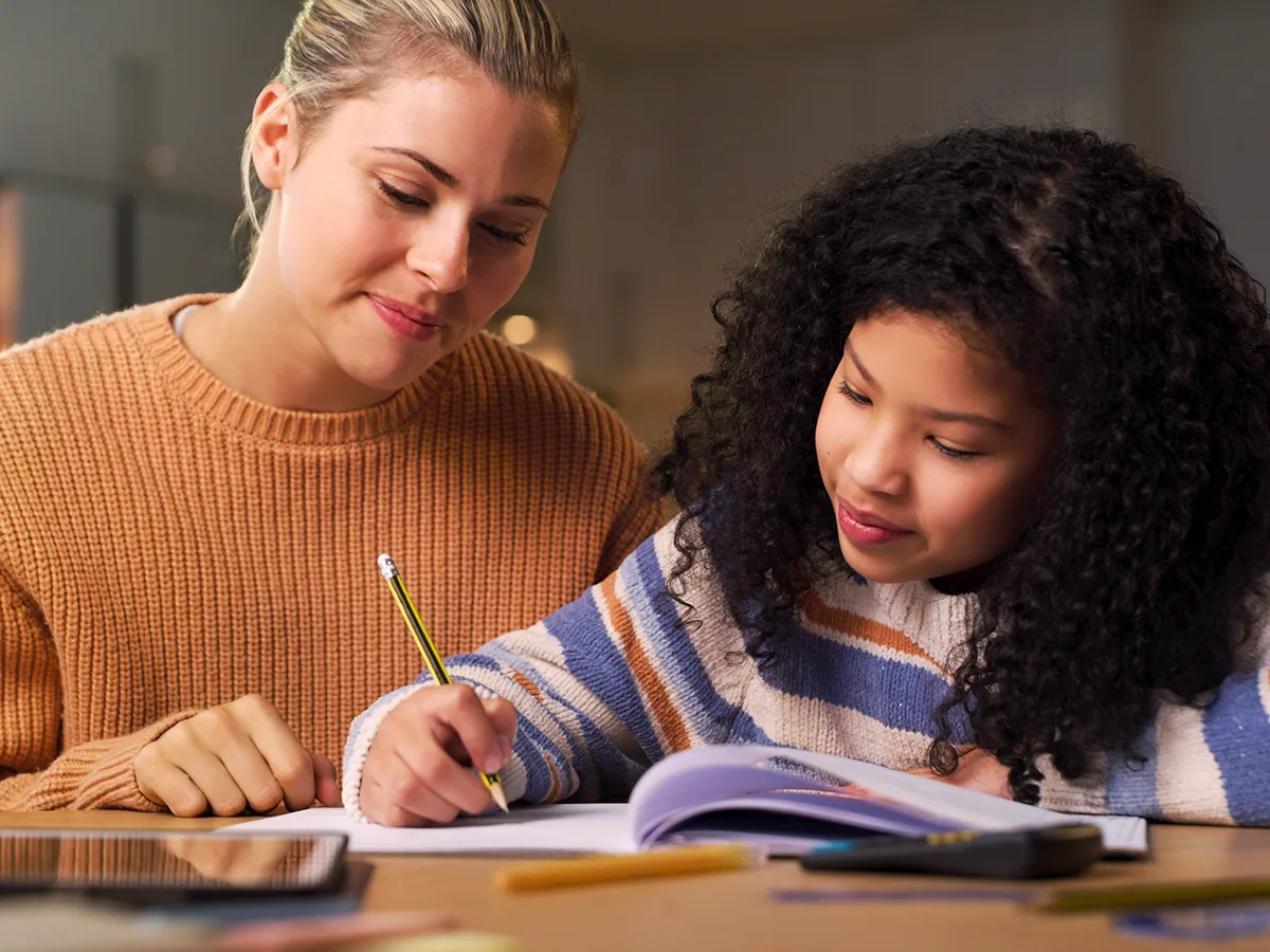 7 ways to boost your child’s vocabulary, black girl sitting at a desk writing in a notebook next to white woman 
