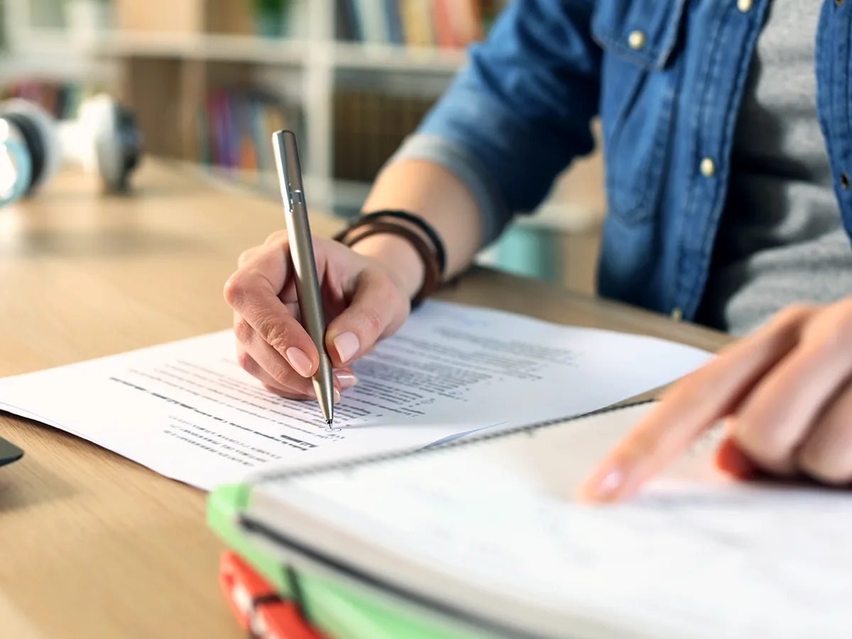 Una adulta señala  un texto en un cuaderno con el dedo índice izquierdo mientras escribe en un documento con su mano derecha.

