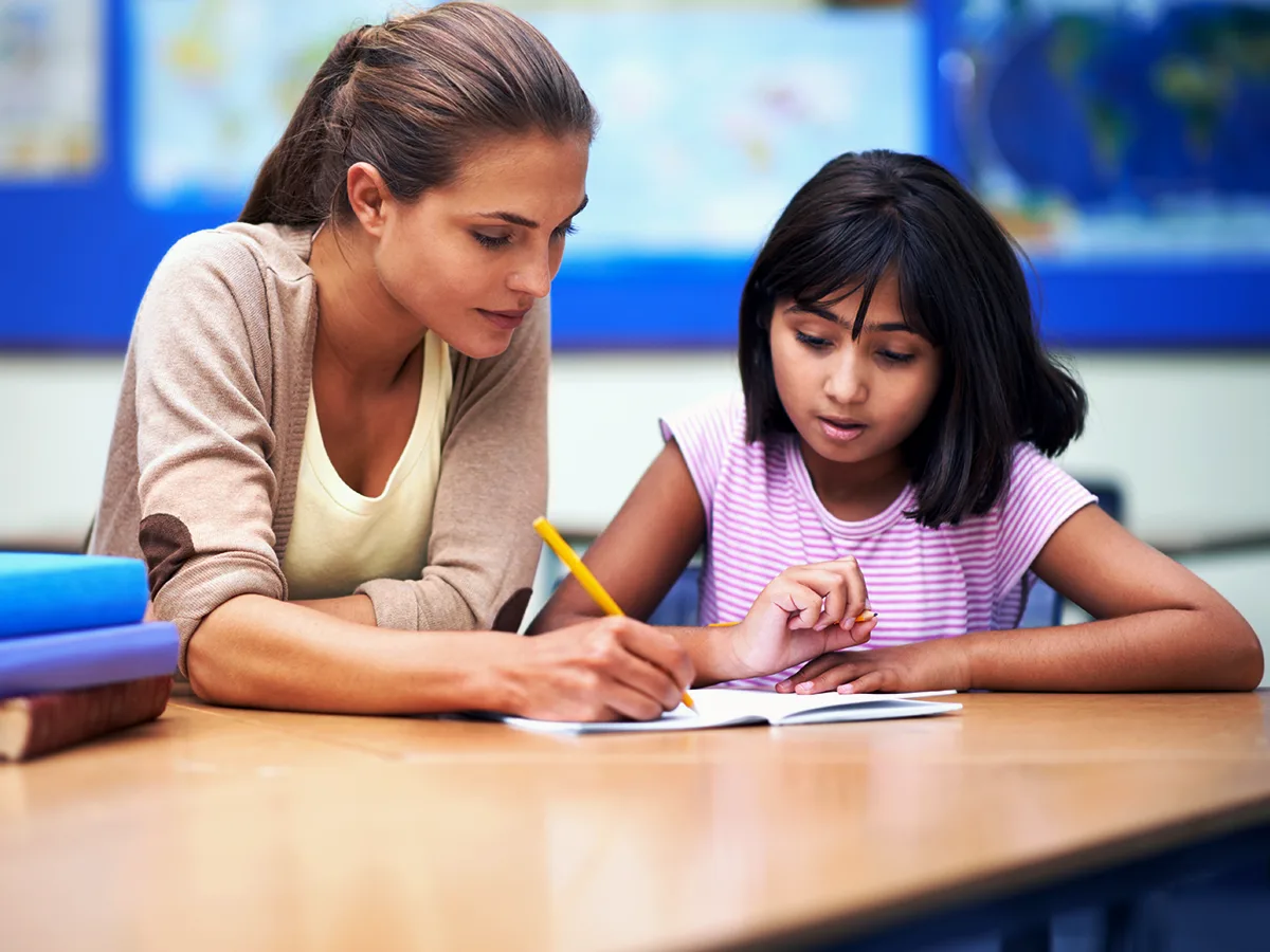 Una maestra muestra la forma correcta de agarrar el lápiz y escribir a una estudiante, quien observa atentamente e imita la posición de los dedos.