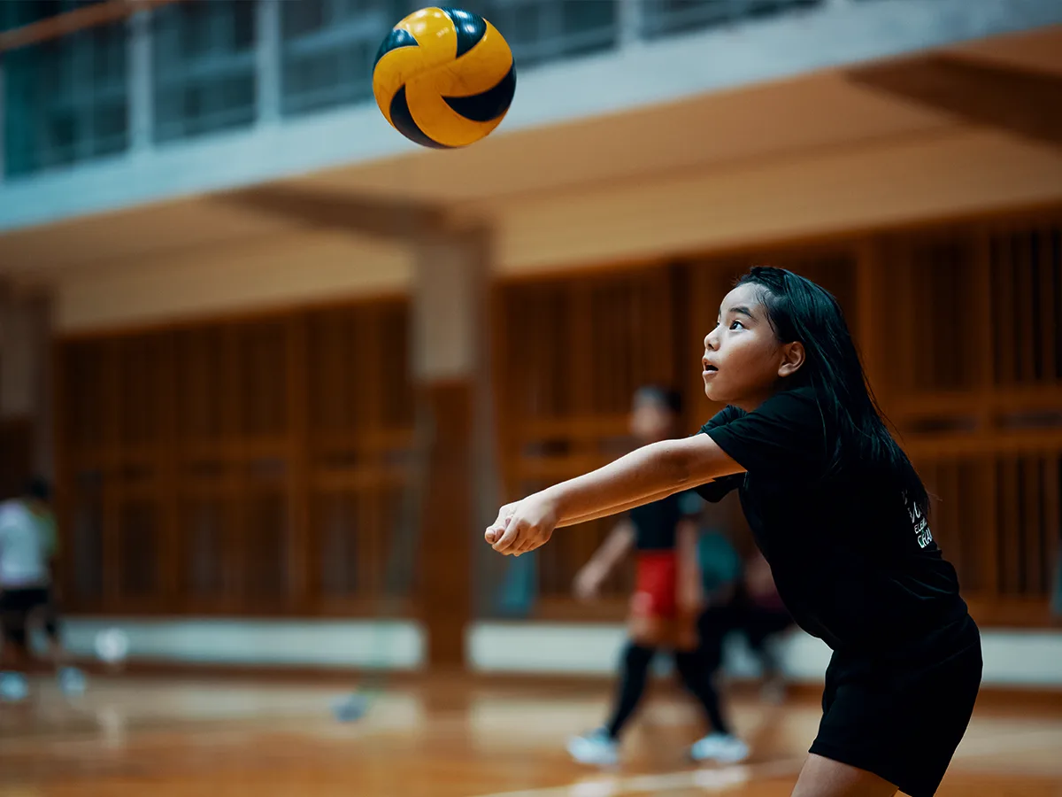 Inside a gymnasium, a child in a black uniform prepares their arms to receive an oncoming volleyball and return it.