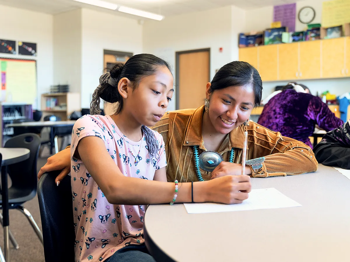 Una maestra está ayudando en el aula a una estudiante con su escritura. La maestra sonríe mientras la estudiante sostiene un lápiz.