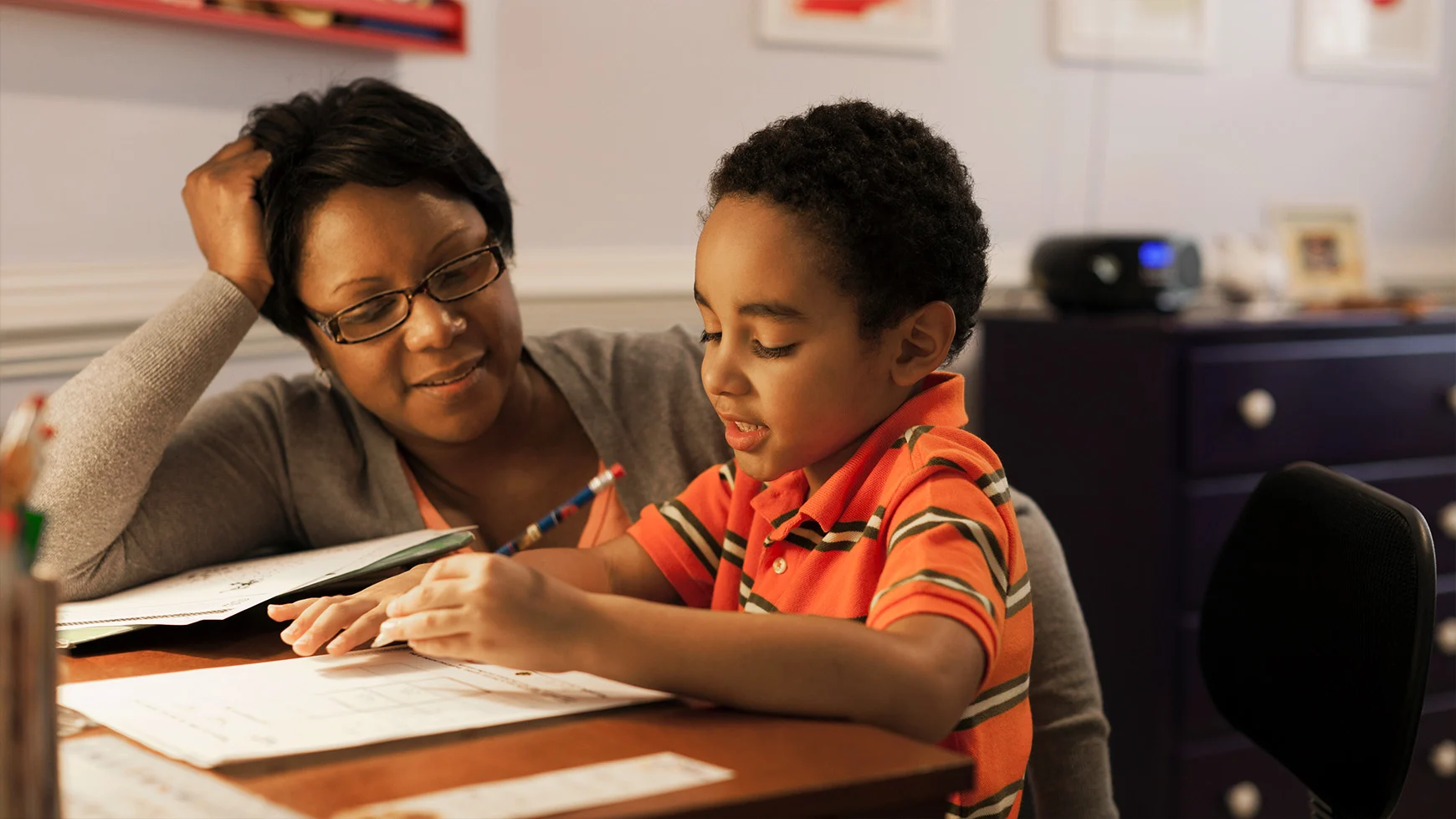 Una mamá sentada con su hijo observa mientras el niño sostiene un lápiz y trabaja en una tarea de escritura. Ambos sonríen.