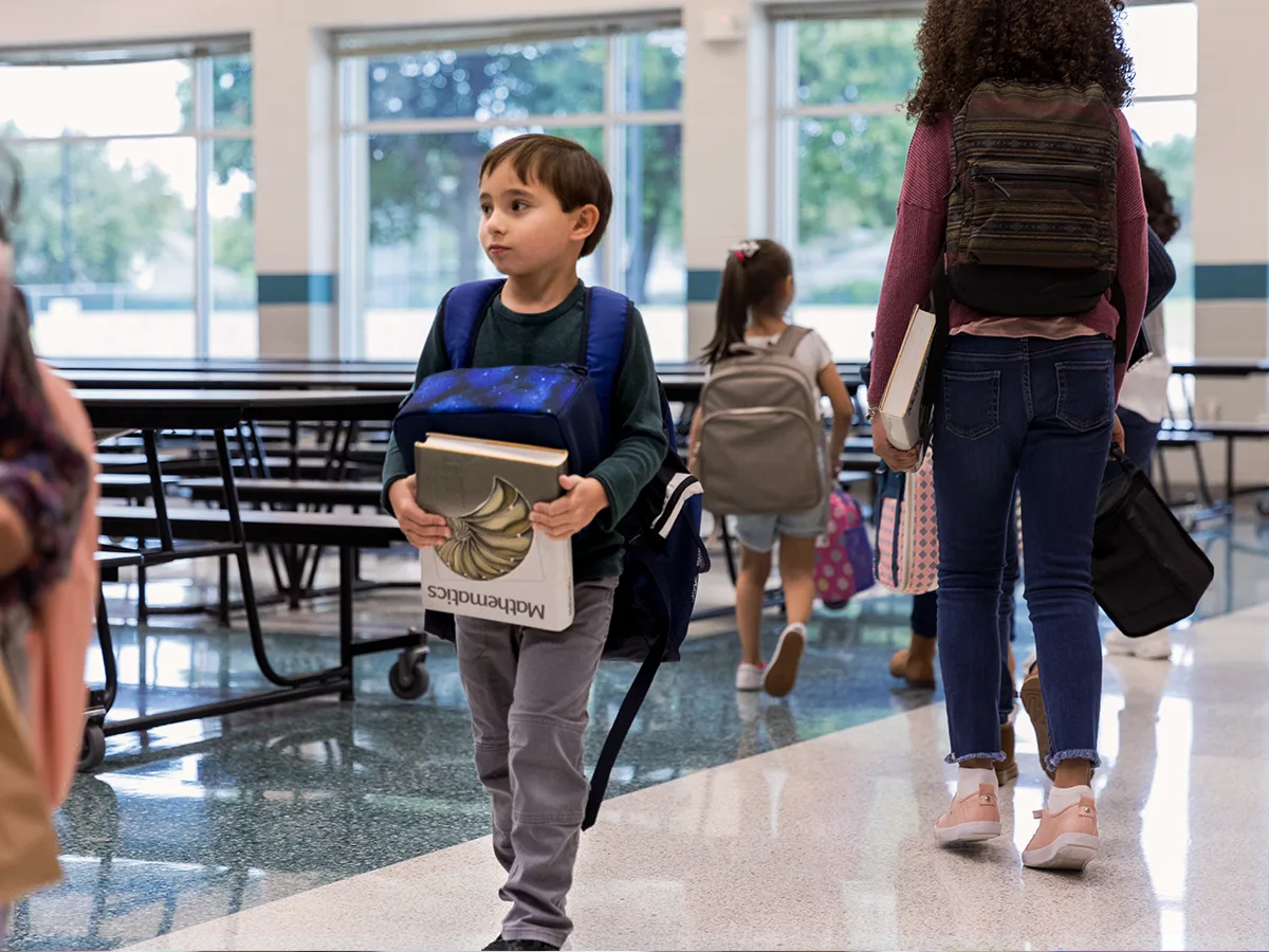 A child walking alone in a cafeteria, holding a mathematics textbook upside down and wearing their backpack on their chest.