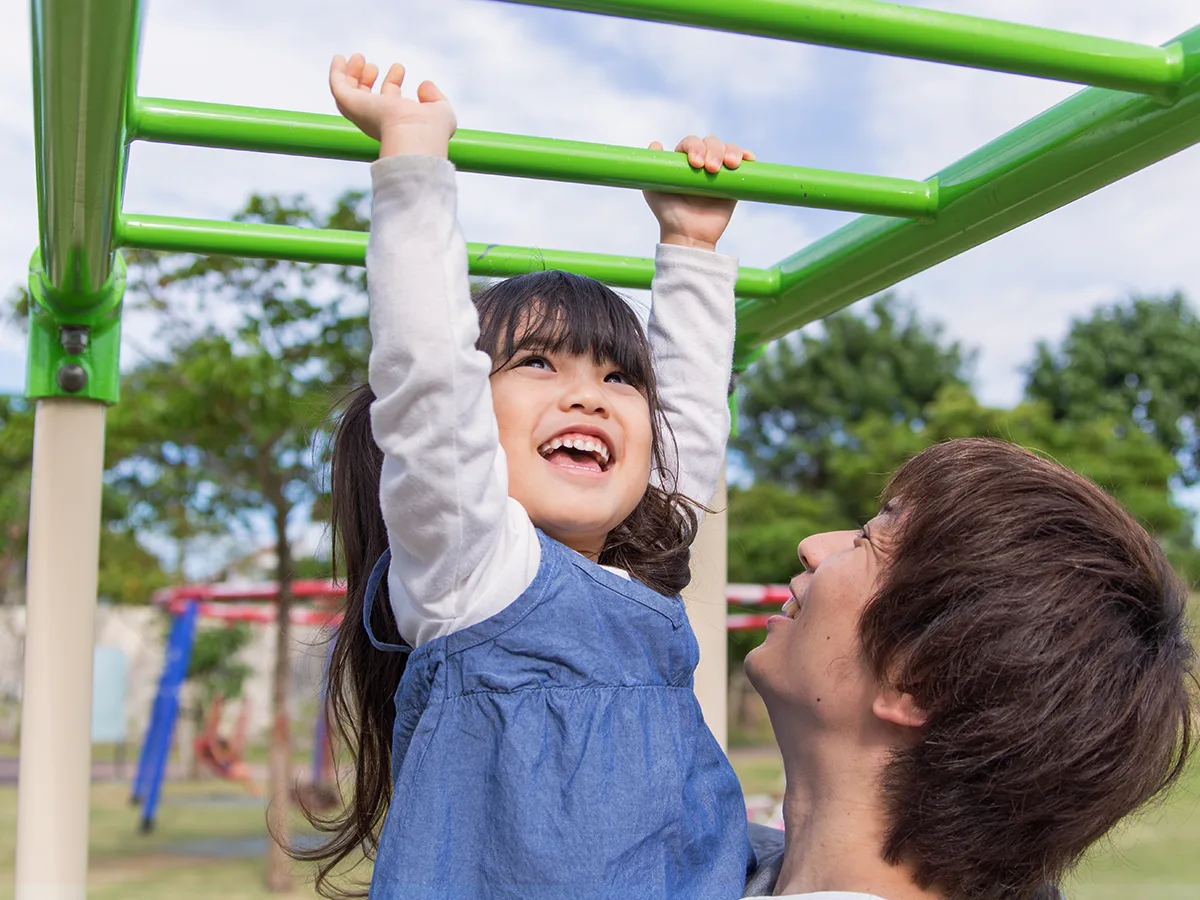 A child hangs on monkey bars on a playground while an adult holds them.