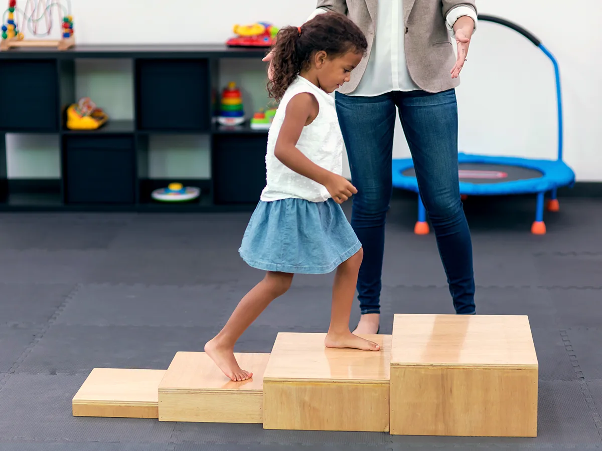 A child climbing a set of stairs in a physical therapy room with an adult standing nearby.