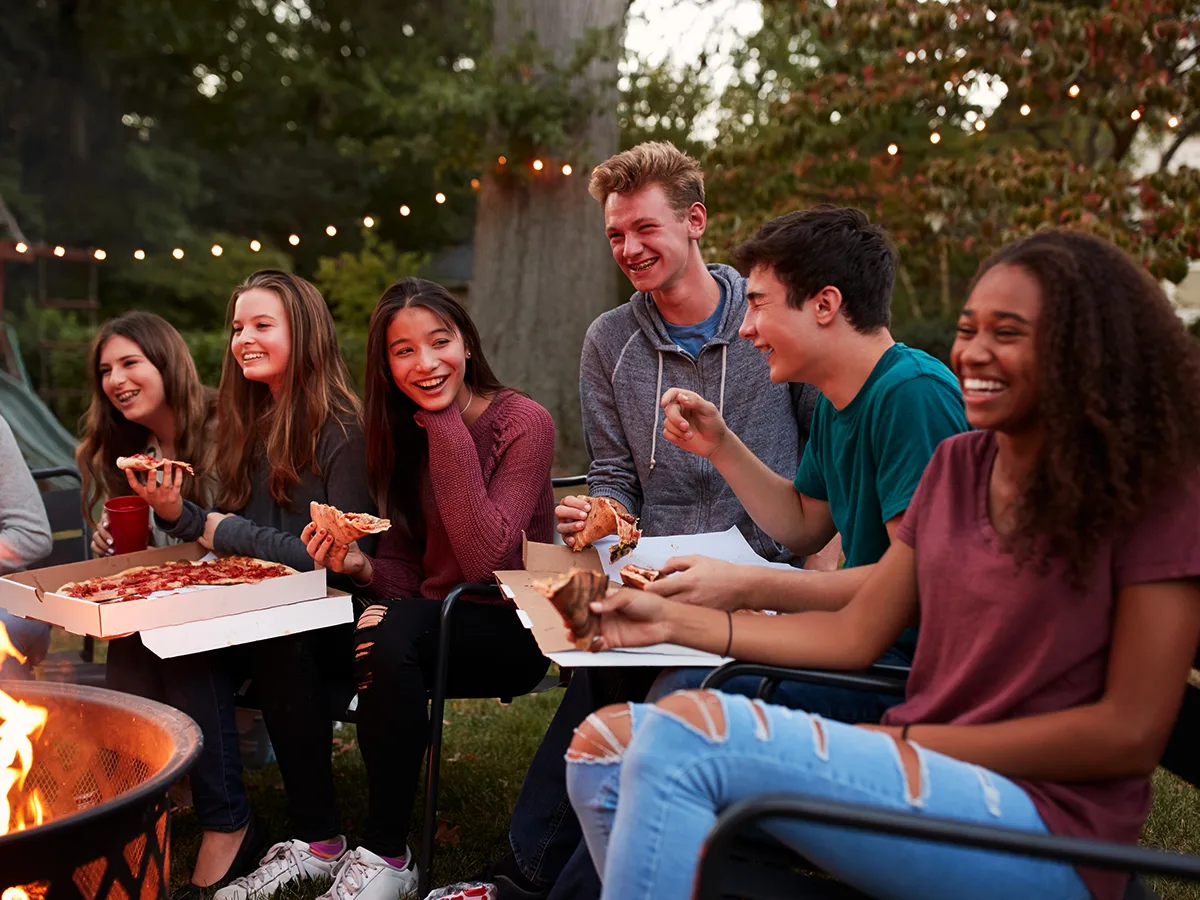 Seis adolescentes sonrientes comen pizza alrededor de un fogón en un jardín al exterior.