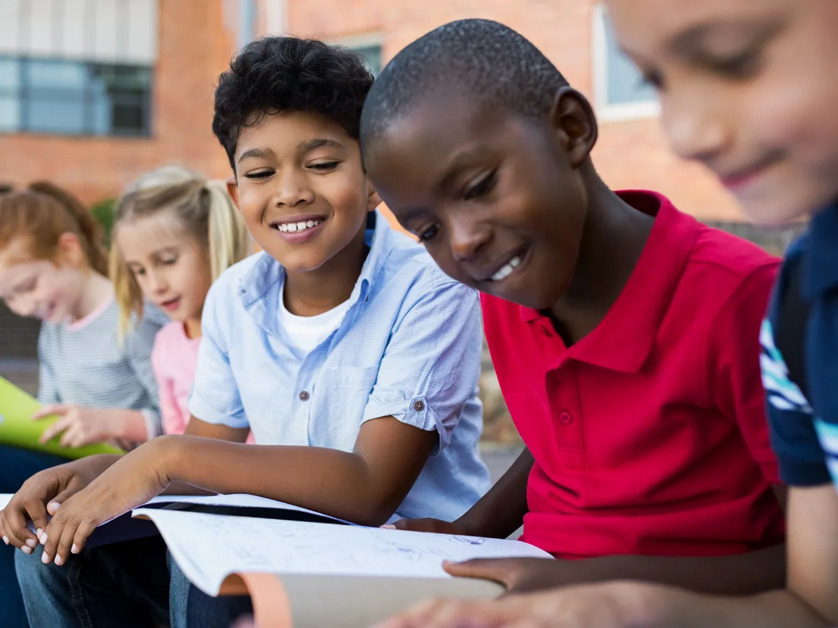 A diverse group of students is reading outside. One student smiles and looks over at another's open notebook.