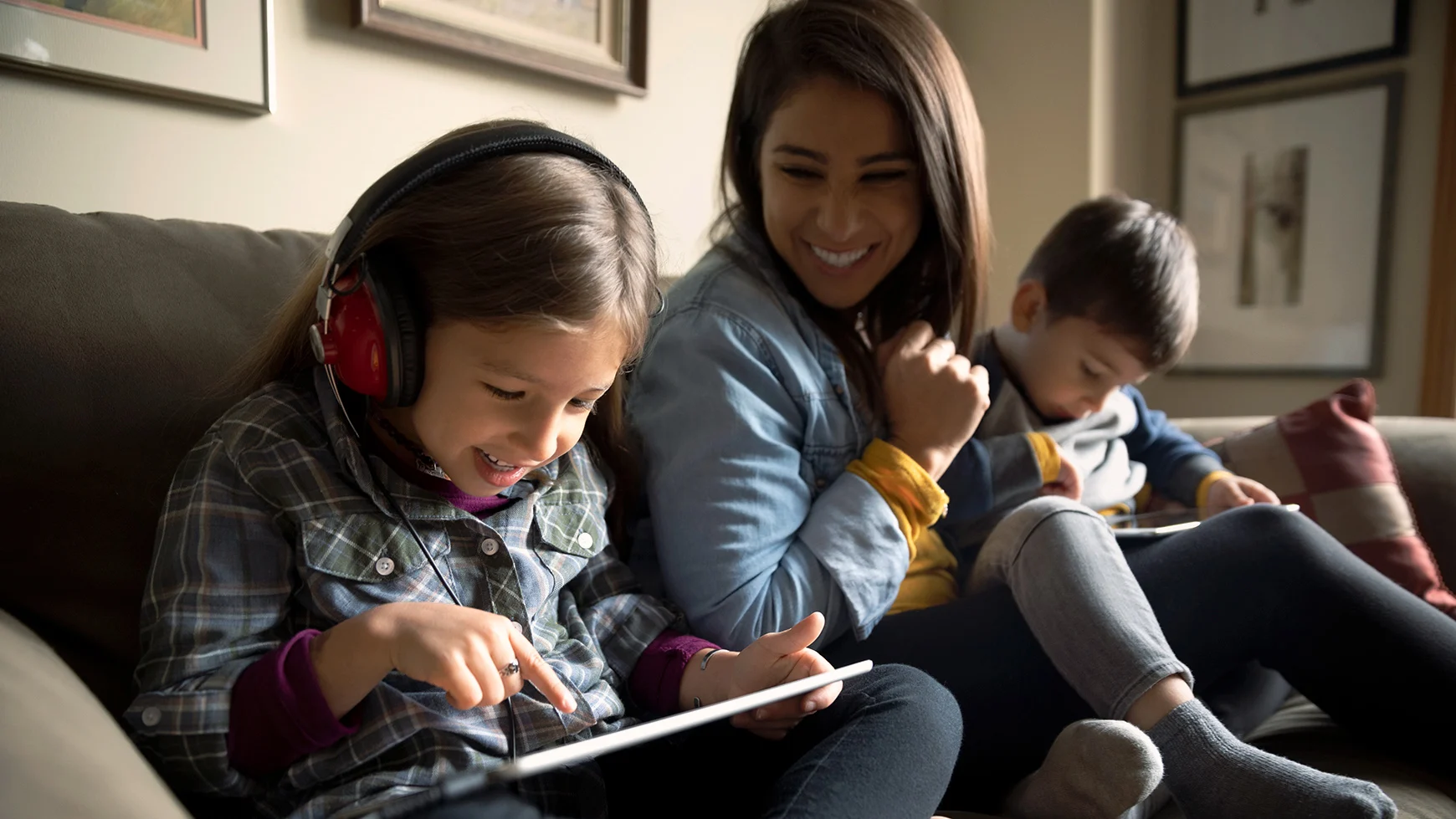 Una adulta y dos niños están sentando en un sofá. Uno de los niños están usando audífonos y ambos niños están utilizando tabletas. 