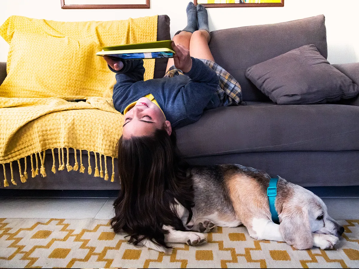 Learning and thinking differences that cause trouble with time management. A girl looks at tablet on couch upside down with her dog. 