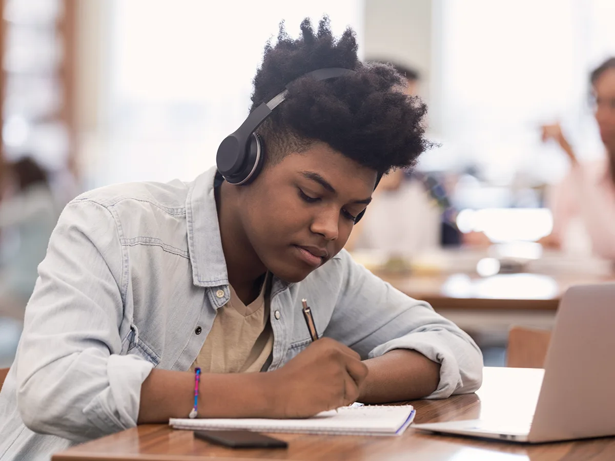 A teen with headphones writes on a piece of paper with a laptop nearby. 