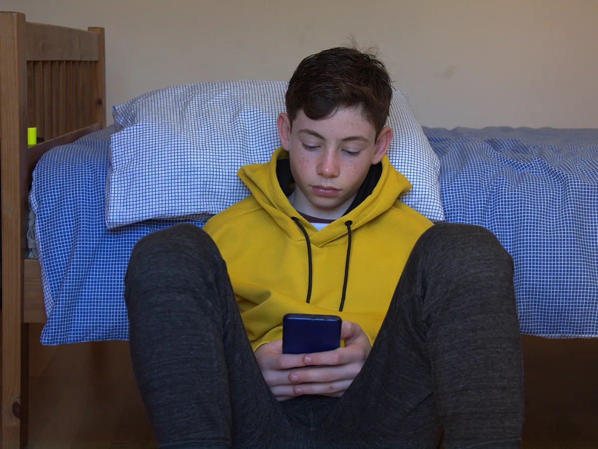 A teen looks at their phone while sitting on the ground leaning against their bed. 