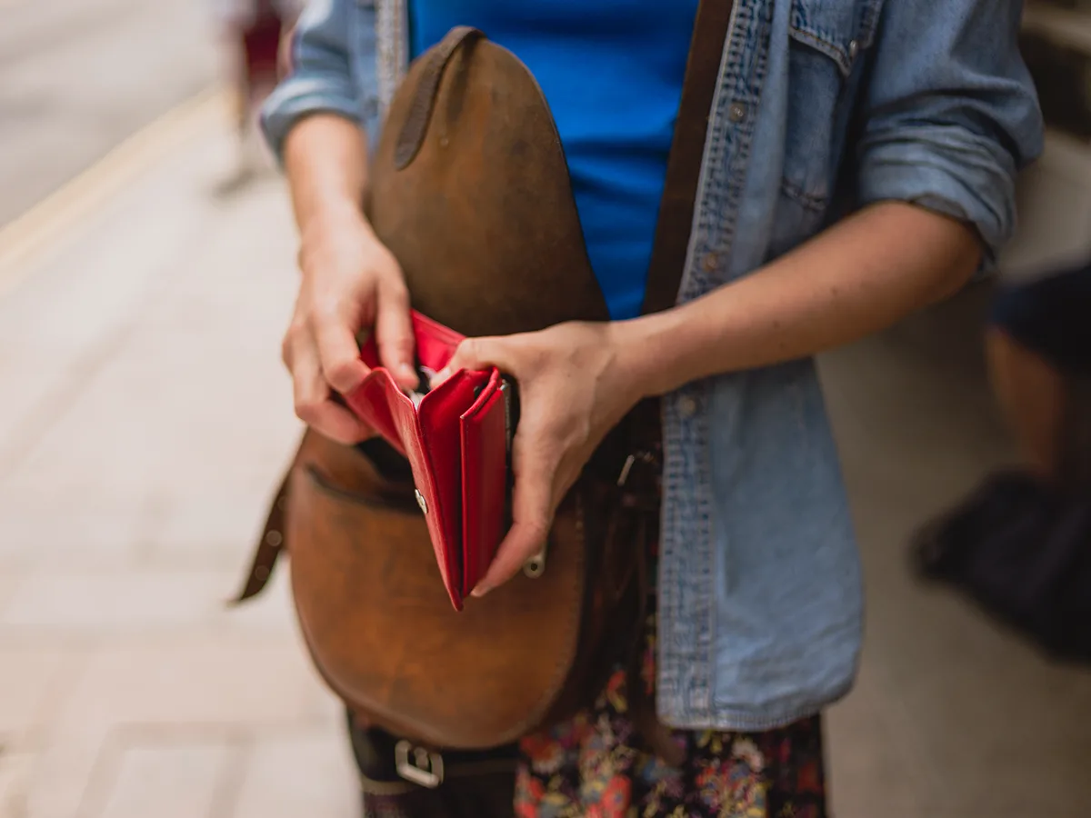 Una persona con un bolso café colgado del hombro sostiene una billetera roja. Está sacando dinero para pagar algo.