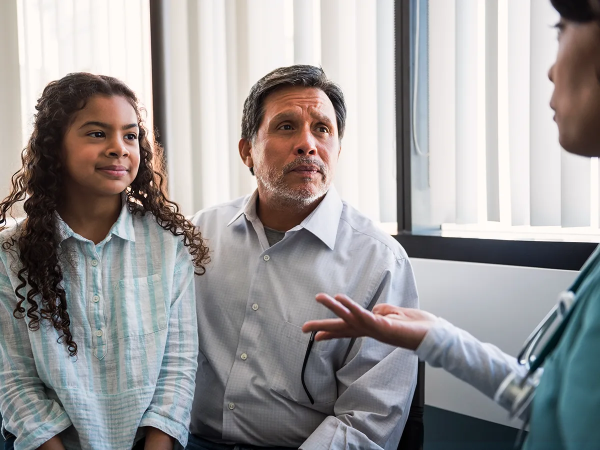 An older adult sits next to a child. The adult talks to a doctor standing nearby. 
