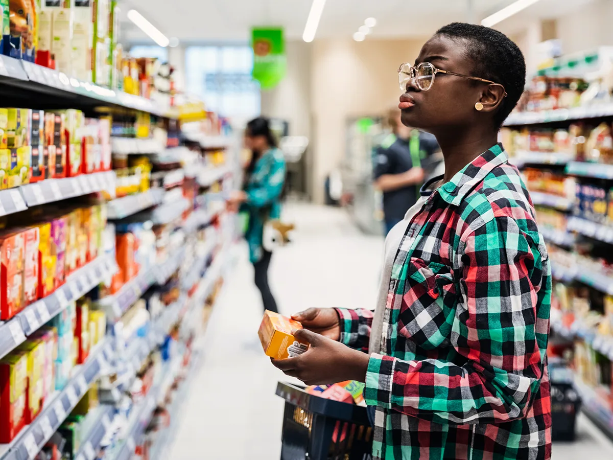 A teenager with a full shopping cart is holding a box in both hands while looking at items on a grocery store shelf.