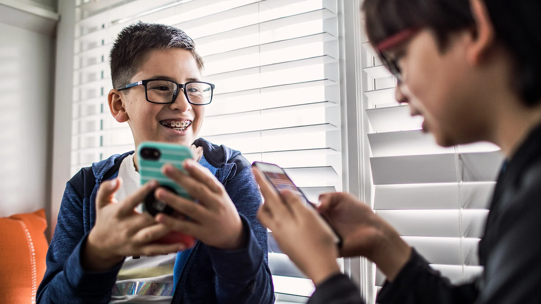 Two kids with glasses are smiling and playing mobile games together. They both hold cell phones. One of them also has braces. 