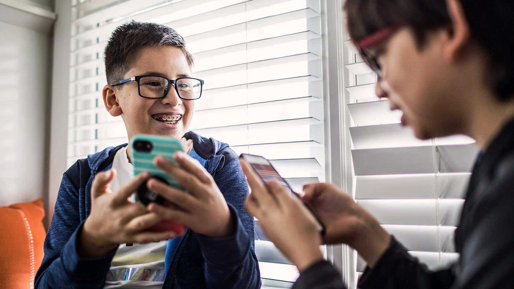 Dos niños con lentes sonríen y juegan videojuegos juntos. Ambos sostienen un teléfono celular. Uno de ellos también usa aparatos de ortodoncia para enderezar sus dientes.