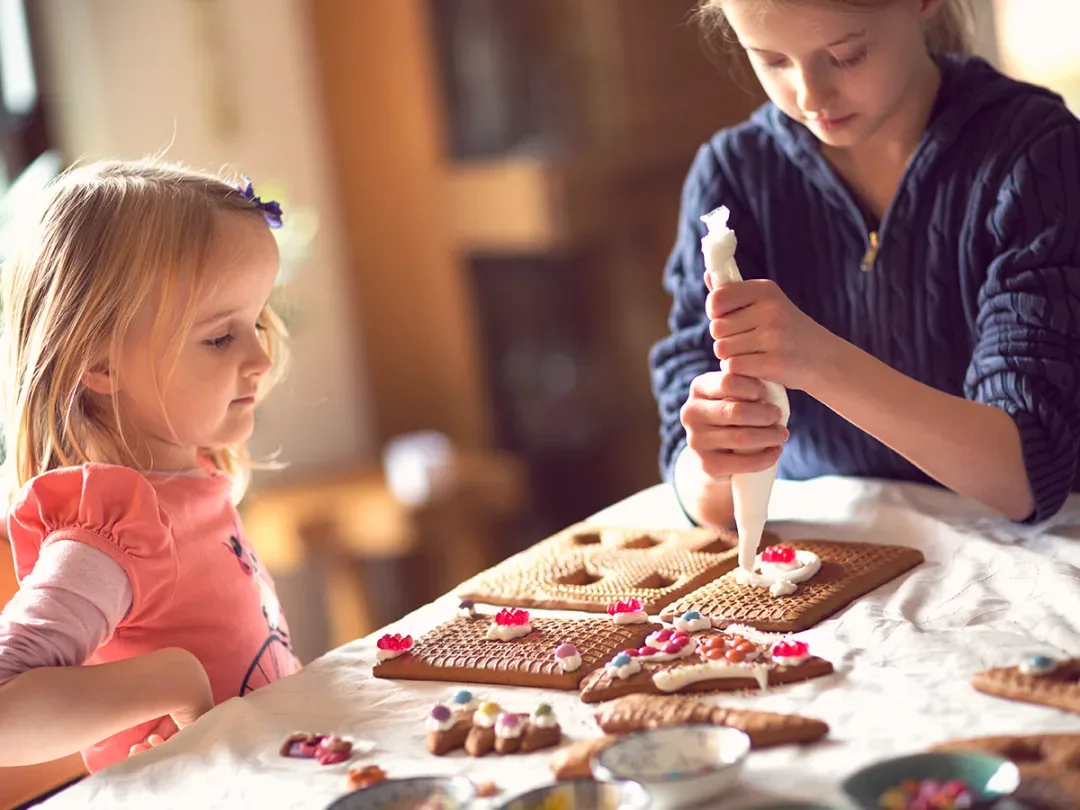Two kids decorate a gingerbread house. One squeezes icing while the other looks at an already-iced piece on the table.