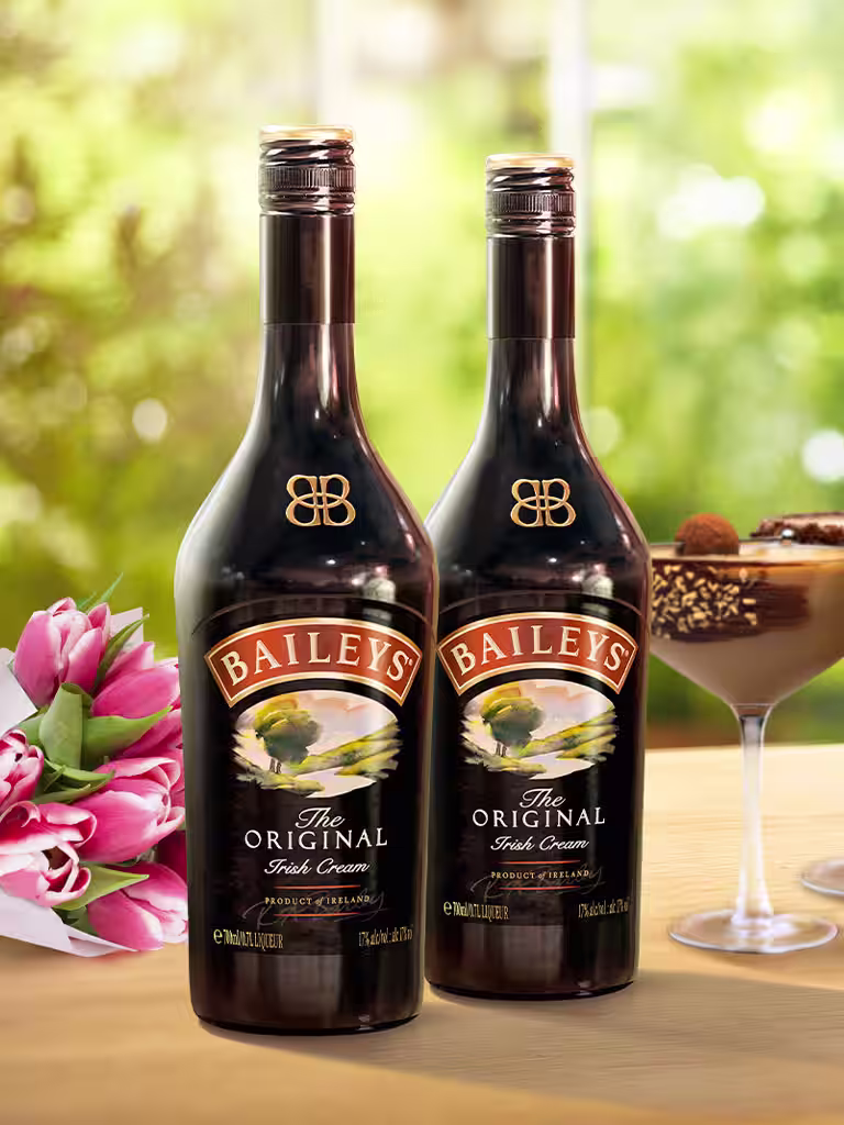 Two Baileys Original Irish Cream bottles on a wooden table next to two martini glasses with chocolaty garnishes, a bouquet of pink tulip.