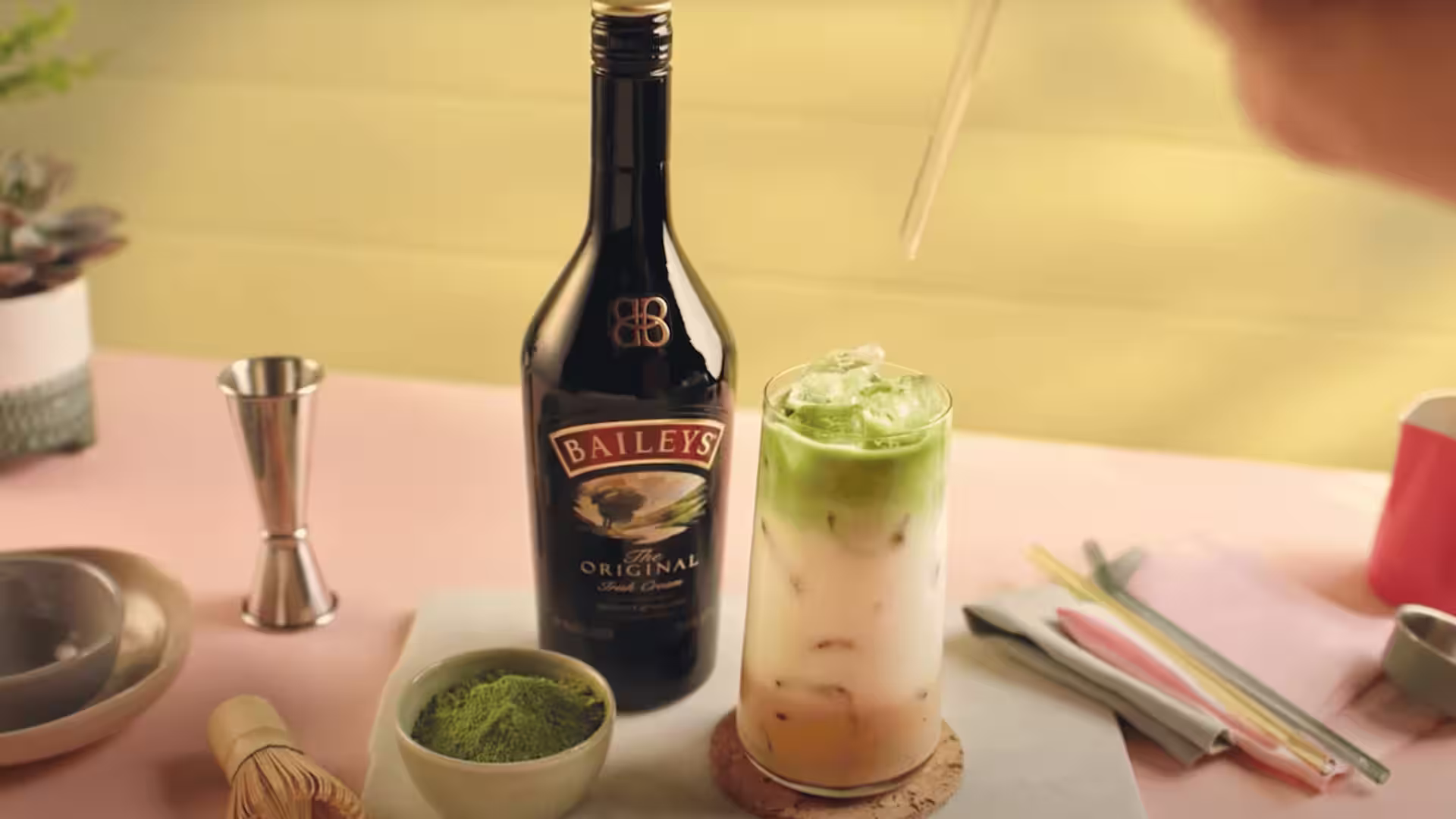 Matcha latte with Baileys, layered with milk and matcha, next to a Baileys bottle, matcha powder, and cocktail tools on a pastel table.