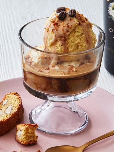 Baileys affogato in a glass dessert dish with vanilla ice cream, espresso, and crumbled toppings, served with biscotti on a pink plate.