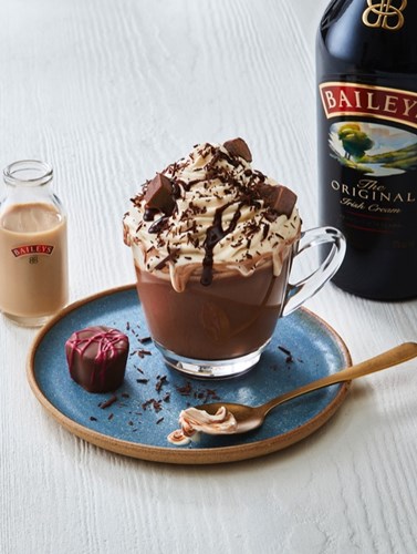 Baileys hot chocolate in a mug, topped with whipped cream, chocolate drizzle, and chunks, served with a truffle on a blue plate, with Baileys bottles.